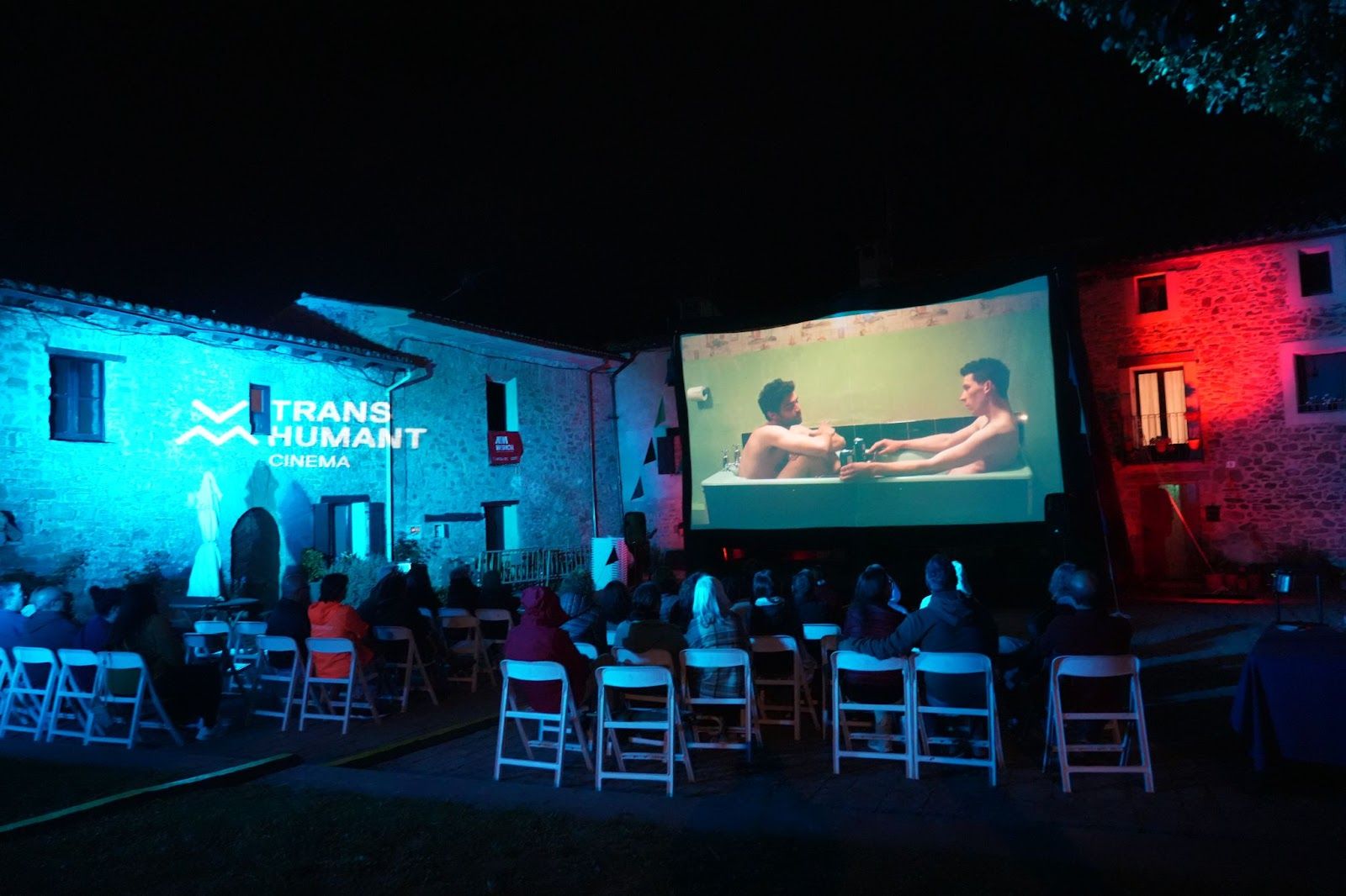 Sessió de cinema a la fresca a la nit en una plaça de Vallfogona del Ripollès. El públic seu en cadires blanques davant una gran pantalla on es projecta una escena amb dos homes. Els edificis de pedra estan il·luminats amb llums blaves i vermelles, i a la paret es llegeix el logotip "TRANS HUMANT CINEMA".