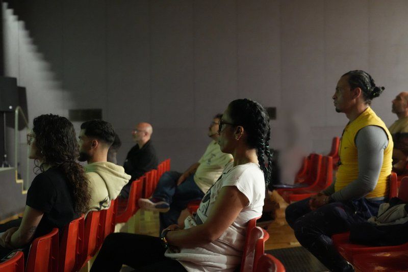 Vista de perfil mitjana d'un grup de persones assegudes en files de cadires vermelles de plàstic en un teatre fosc o sala d'espectacles. Totes les figures estan mirant cap a l'esquerra, en direcció a un espai fosc de fons, possiblement un escenari o pantalla. En primer pla, a l'esquerra, hi ha una dona amb els cabells llargs, foscos i arrissats, que porta una samarreta blanca amb un gràfic i està asseguda de perfil. Més a la dreta, hi ha un home amb els cabells recollits, que porta una armilla groga brillant sobre una samarreta grisa de màniga llarga, assegut de perfil. Al fons, es poden veure altres persones assegudes, inclosa una dona amb tresses i ulleres de sol, i un home amb una dessuadora beige amb caputxa. L'enfocament és nítid en les dues figures principals en primer pla i es difumina cap al fons.
